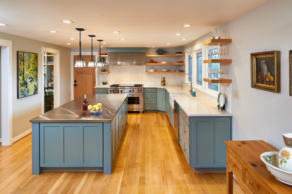 rustic modern kitchen with blue gray cabinets that highlight one of our three key interior design trends in 2021, open shelving and lots of counterspace. Island has stainless steel countertop and flooring is a natural light hardwood