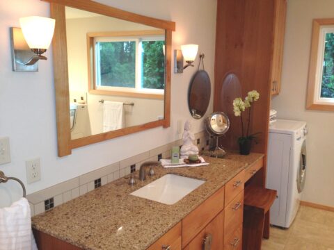 Accessible bathroom vanity using universal design. Brown speckled countertop with large vanity mirror and dual combination washer and dryer. In mirror's reflection is a window wheelchair accessible bath tub.