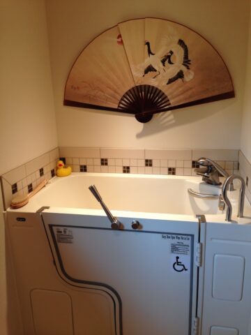 Wheelchair accessible bath tub created with universal design. Yellow rubber ducky in the corner of tub and Asian hand fan artwork on the wall. 